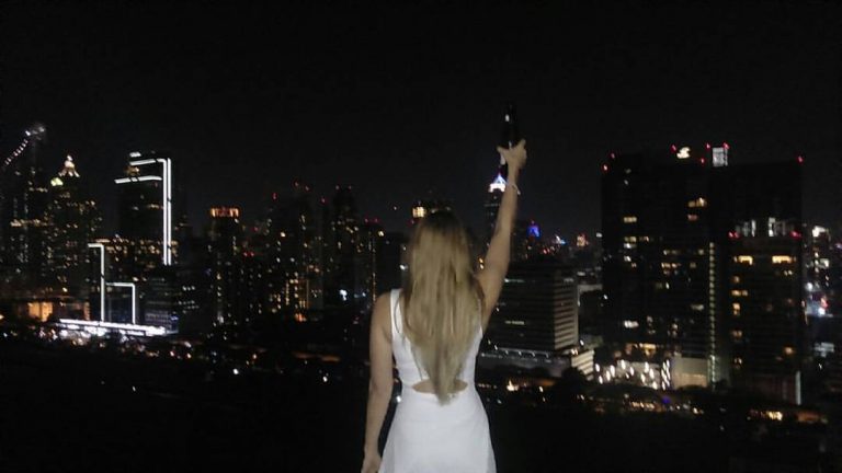 Steffi, a Bangkok based travel blogger stands in a white dress holding a beer bottle to the sky on the top of a rooftop at night overlooking the lights of Bangkok