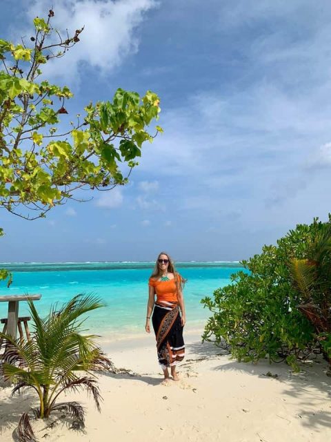 Steffi a travel blogging working in the Maldives wears a black and orange traditional Maldivian sarong and an orange top standing on a white sand beach with greenery around and a turquoise sea behind with cloudy skies