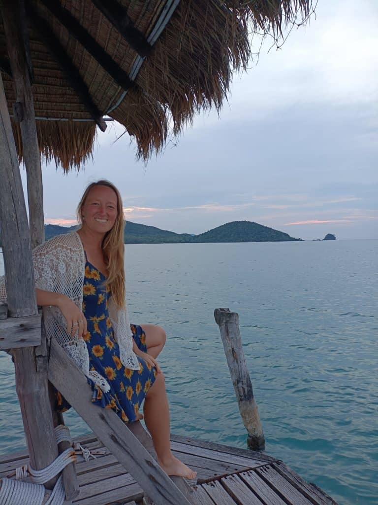 steffi, a teacher in thailand, white blonde sits on a pier looking at the camera with the thai sea in the background and a hut with fringed coconut palms at the top