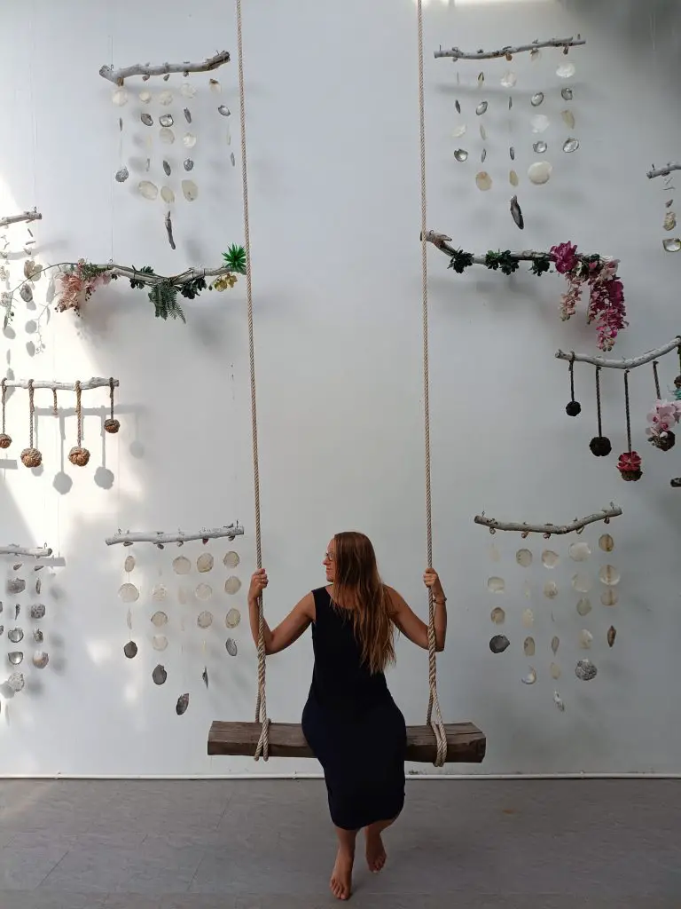 Steffi, a UK teacher working in Bangkok, sits on a swing in a fancy resort in Thailand. The background is a white wall with shell decorations