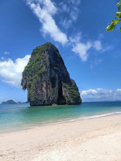 A huge limestone karsts sticks out of the turquoise water with a white sand beach in the foreground in Railay Thailand. A blue sky with 3 little fluffy clouds is in the background