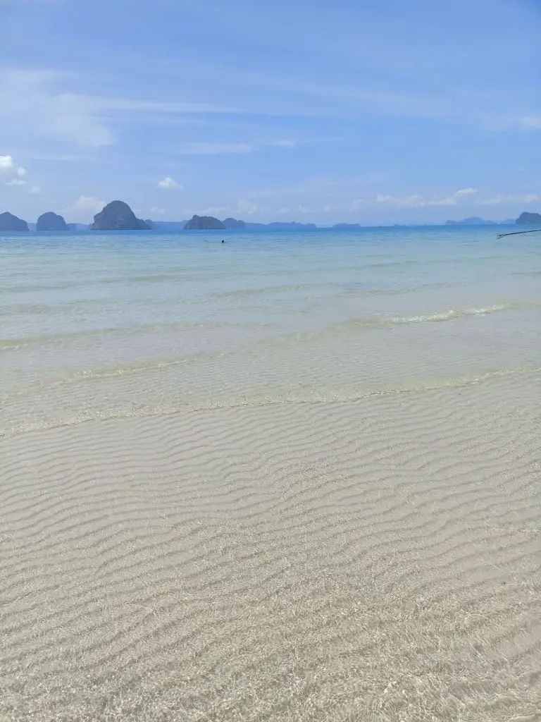white sand beach in thailand with clear water and phangna bay limestone karsts in the background and a blue sky