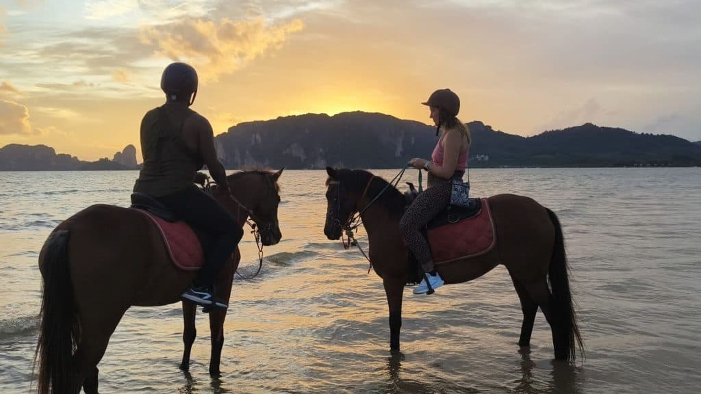 Jason and Steffi, two expats living in Bangkok, horse riding on the beach in Krabi at sunset. They are silhouetted with sunset over the mountains in the background